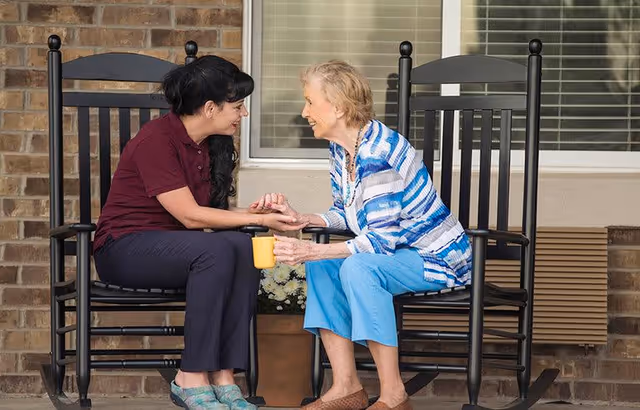 An elderly woman and a younger woman sitting on black rocking chairs on a porch, holding hands and smiling at each other. The elderly woman is holding a yellow mug, and there is a window with blinds and a brick wall behind them.