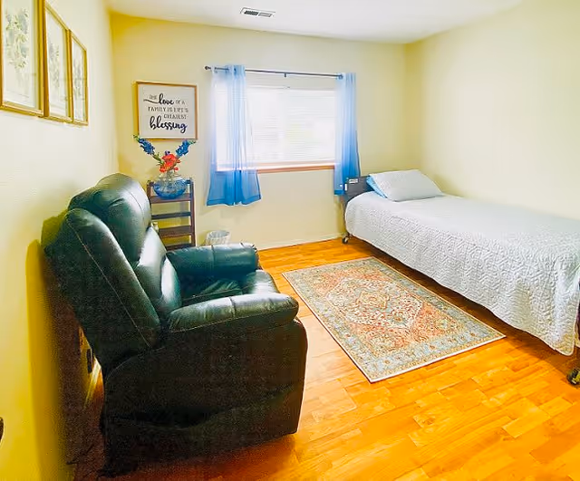 A small, cozy bedroom with a single bed covered in a white quilt, a black leather recliner chair, a colorful area rug on a wooden floor, a window with blue curtains, and a small table with a vase of flowers and a framed inspirational quote on the wall.