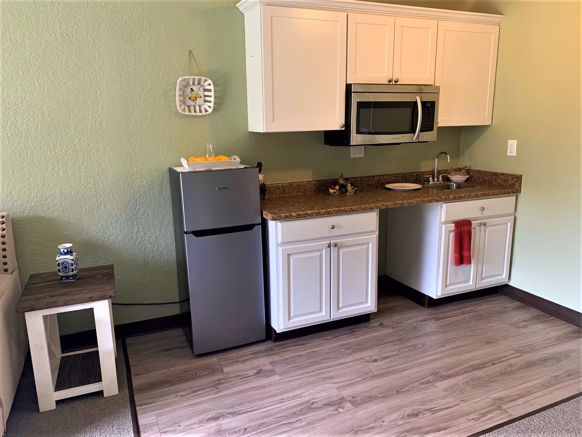 A small kitchen area with white cabinets, a brown countertop, a stainless steel microwave mounted above the counter, and a compact refrigerator. There is a small sink with a faucet, a red towel hanging on one cabinet door, and decorative items on the countertop. The walls are painted light green, and there is a small wooden side table with a blue and white vase next to a beige couch.