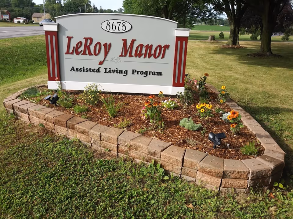 A landscaped sign for Leroy Manor Assisted Living Program with the address number 8678 displayed above the name. The sign is surrounded by a small garden bed with flowers and mulch, bordered by stone bricks. Trees and grass are visible in the background.