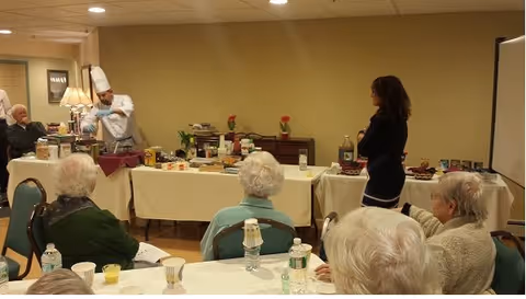 A group of elderly people seated around tables in a room, watching a chef in a white uniform and hat demonstrating or preparing food behind a table covered with various food items. A woman stands nearby observing the chef. The setting appears to be a community or activity room.