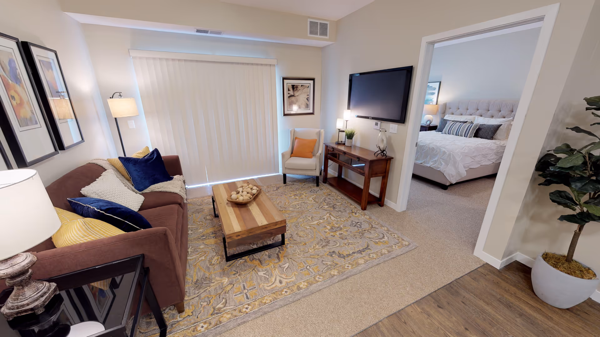 Well-lit living room with a sofa, coffee table, area rug, TV on a console and an adjacent bedroom visible through an open doorway.