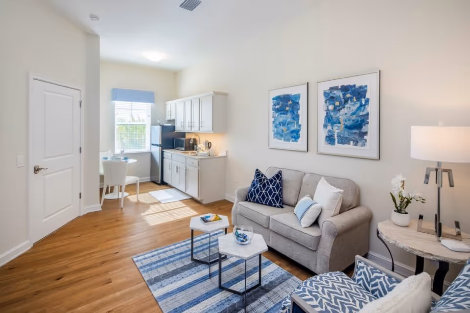 Bright furnished living room with a small kitchenette, gray sofa, patterned chairs, coffee tables, and blue artwork on the wall.