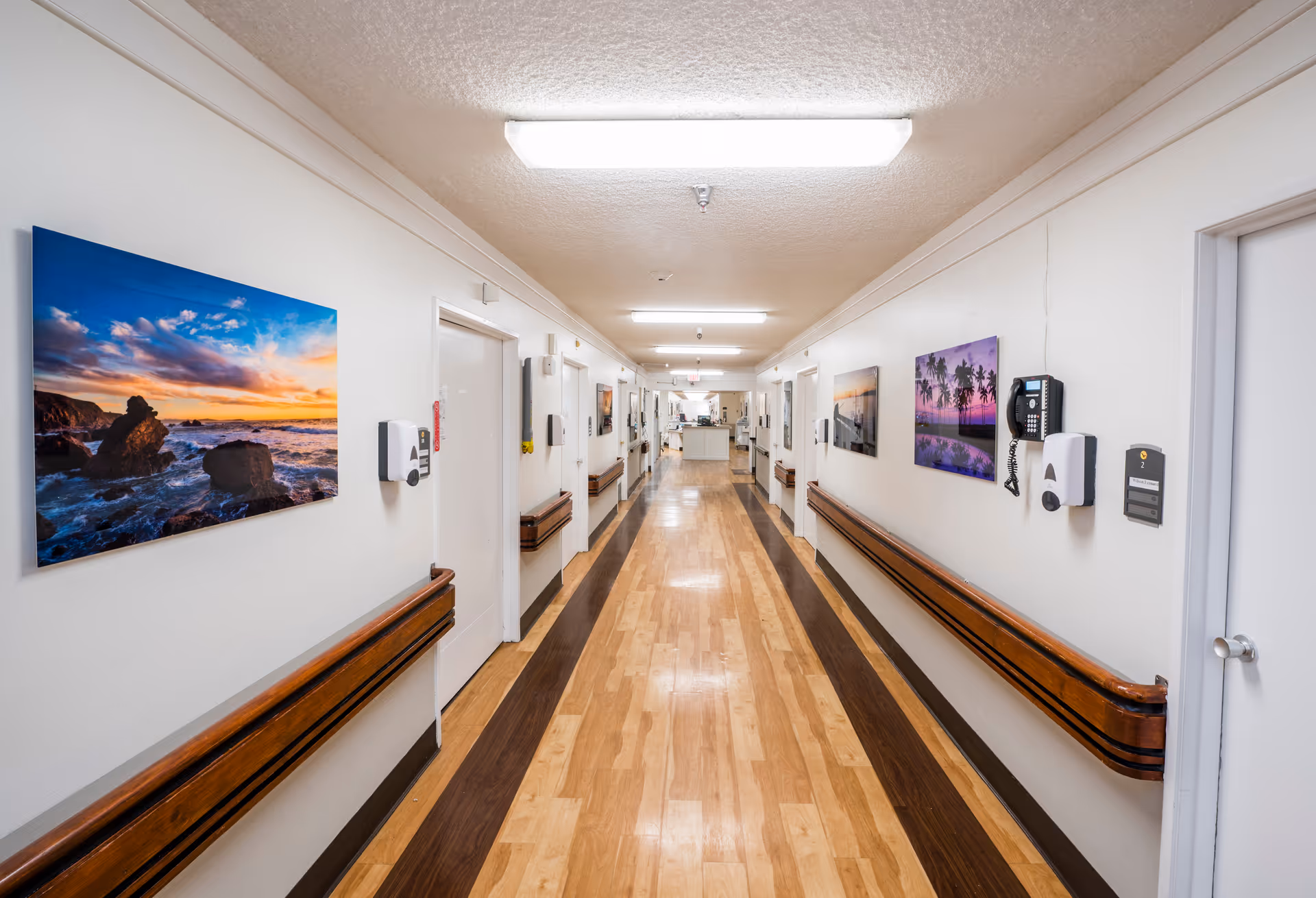 A long, well-lit hallway in a senior living facility with wooden handrails on both sides and several closed doors. The walls are decorated with colorful landscape photographs, and there are wall-mounted hand sanitizer dispensers and a telephone. The floor has a light wood pattern with darker wood borders.