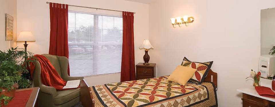 A cozy bedroom with a single bed covered in a colorful quilt and two pillows. Next to the bed is a wooden nightstand with a lamp. A green armchair with a red throw blanket and an open book sits near a window with red curtains and white blinds. There are plants on a dresser and a countertop with a soap dispenser and a small flower arrangement.