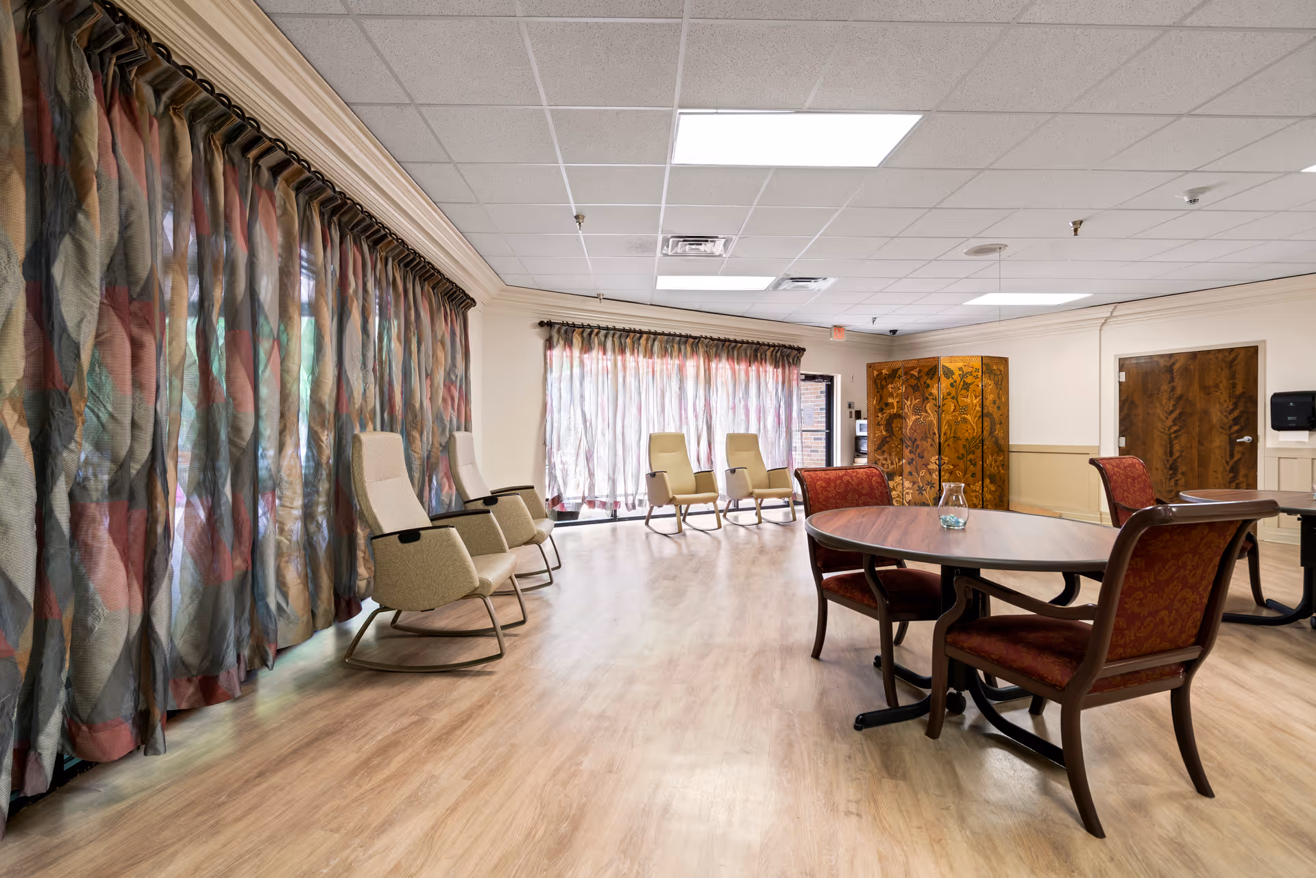 A spacious room with large windows covered by patterned curtains, several beige rocking chairs lined up along the windows, and round wooden tables with red upholstered chairs. The room has a light wood floor and a white drop ceiling with fluorescent lights.