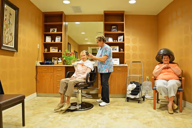 A senior woman is seated in a salon chair having her hair styled by a caregiver in a cozy room with warm yellow walls and wooden cabinets. Another senior woman sits nearby under a hair dryer, wearing an orange top and light pants. The room has a tiled floor, shelves with framed photos, and a floral painting on the wall.