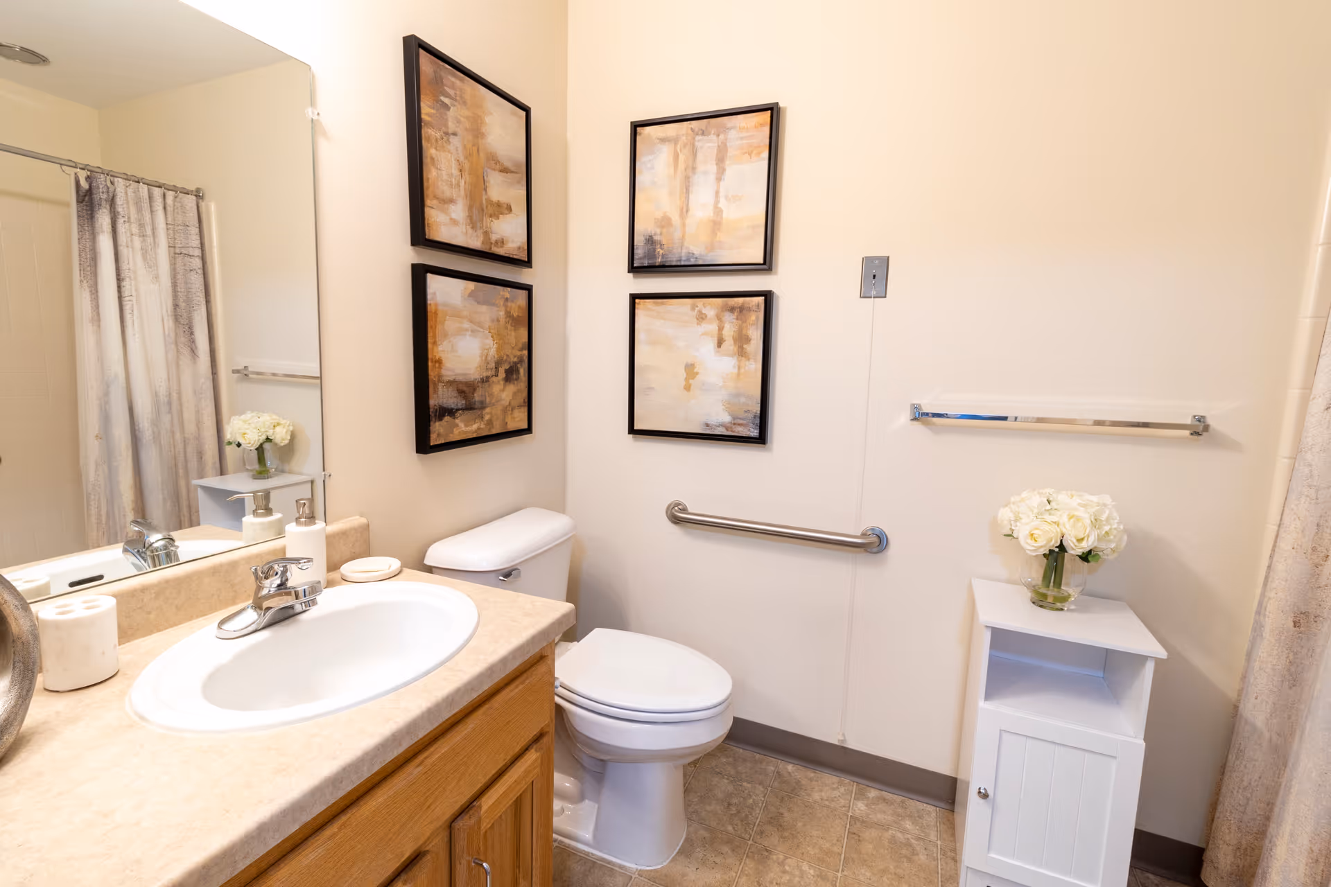 A clean bathroom featuring a sink with a beige countertop and wooden cabinet, a toilet with a grab bar on the wall beside it, three abstract paintings on the wall, a small white cabinet with a vase of white flowers, a towel rack, and a shower with a curtain.