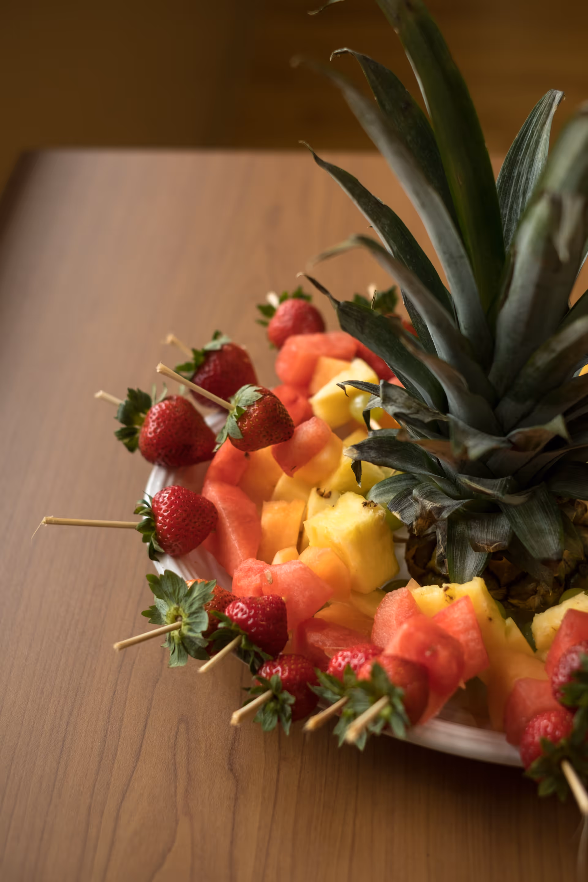 A platter of fresh fruit arranged around the top of a pineapple, including skewers with strawberries, watermelon, and pineapple chunks on a wooden table.