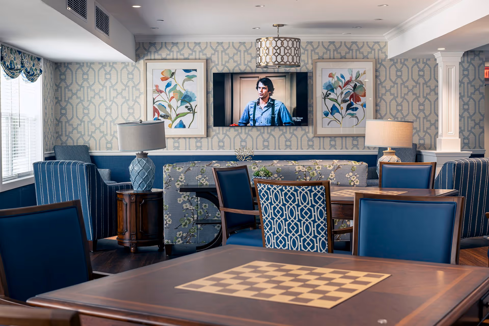 A cozy common area with blue and floral patterned chairs and sofas arranged around wooden tables, one of which has a built-in chessboard. The walls feature geometric patterned wallpaper with two framed floral paintings and a flat-screen TV mounted between them. Two table lamps with blue bases and beige shades provide additional lighting.