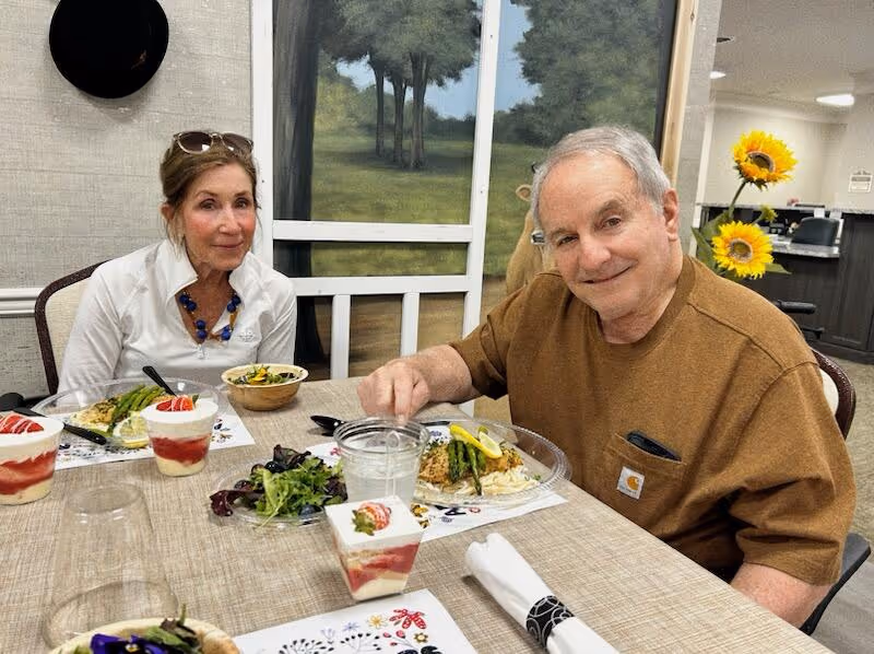 An elderly man and woman sitting at a dining table with plates of food and desserts in front of them. The man is smiling and wearing a brown shirt, while the woman is wearing a white shirt and a blue necklace. Behind them is a wall with a painting of trees and sunflowers in a vase on a counter.