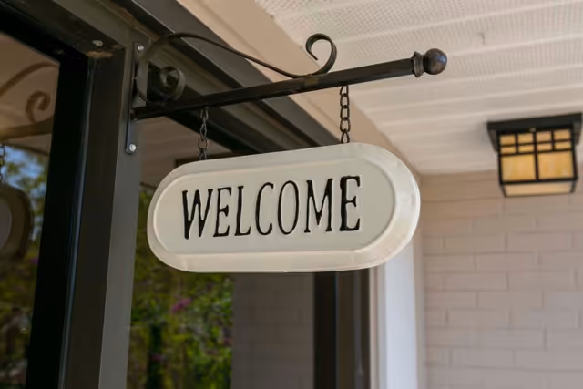 A white hanging sign with the word 'WELCOME' in black letters, mounted on a black metal bracket attached to the ceiling of a porch area. The background shows part of a brick wall painted light gray and a black-framed glass door.