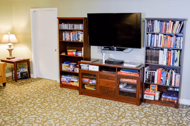 A cozy living room area with a patterned carpet, a wooden entertainment center holding a flat-screen TV, board games, DVDs, and bookshelves filled with books and games. A side table with a lamp and a chair are visible on the left side.