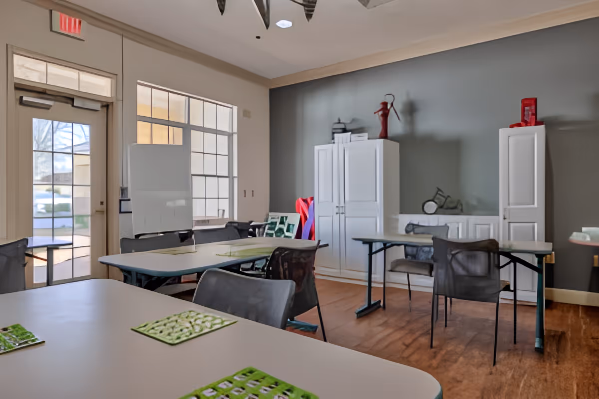 A well-lit room with several tables and chairs arranged for group activities or meetings. The room has large windows and a glass door letting in natural light. There are white cabinets against a gray wall, with decorative items on top. The floor is wooden, and the overall atmosphere is clean and organized.