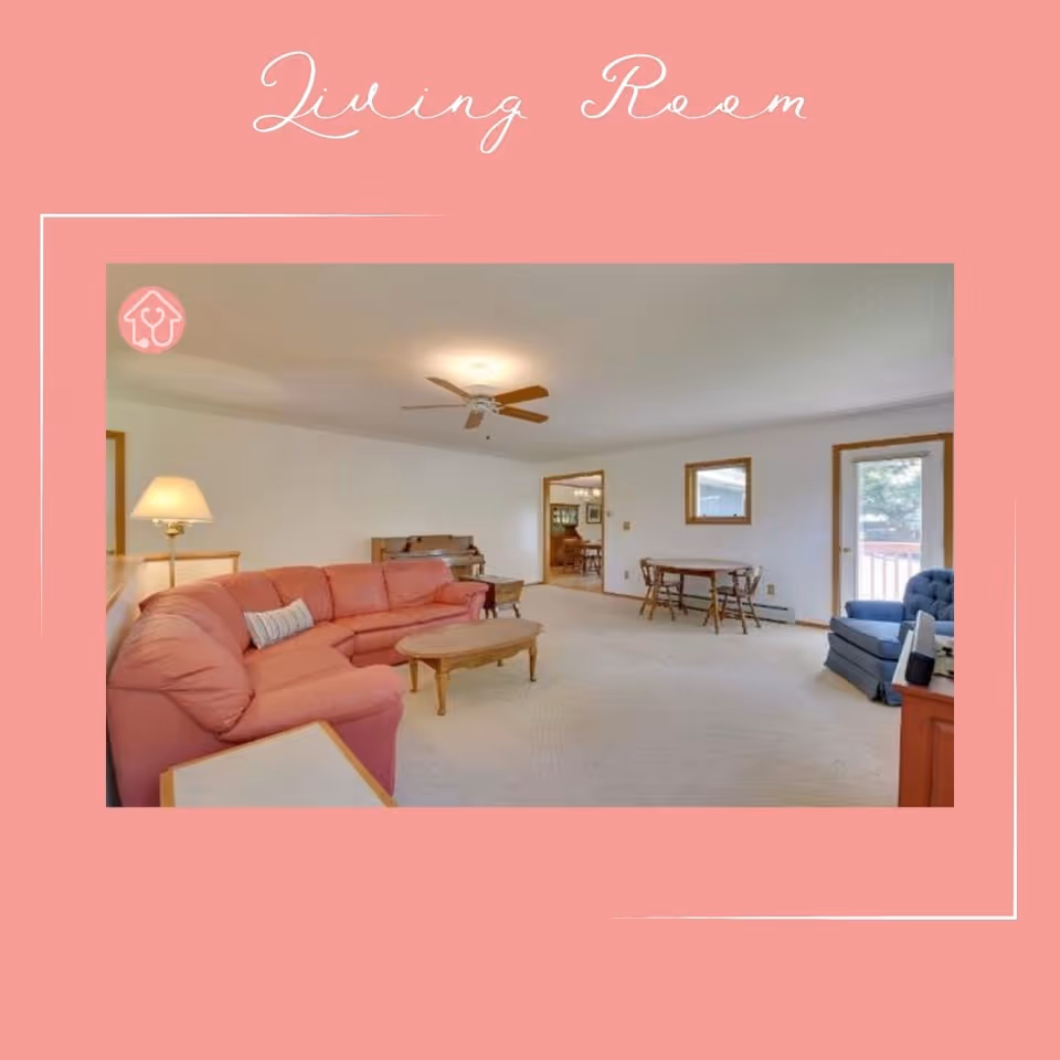 A spacious living room with a pink sectional sofa, a wooden coffee table, a blue armchair, a small dining table with chairs, a ceiling fan, a floor lamp, and a door leading outside. The walls are white and there is a mirror hanging on one wall.