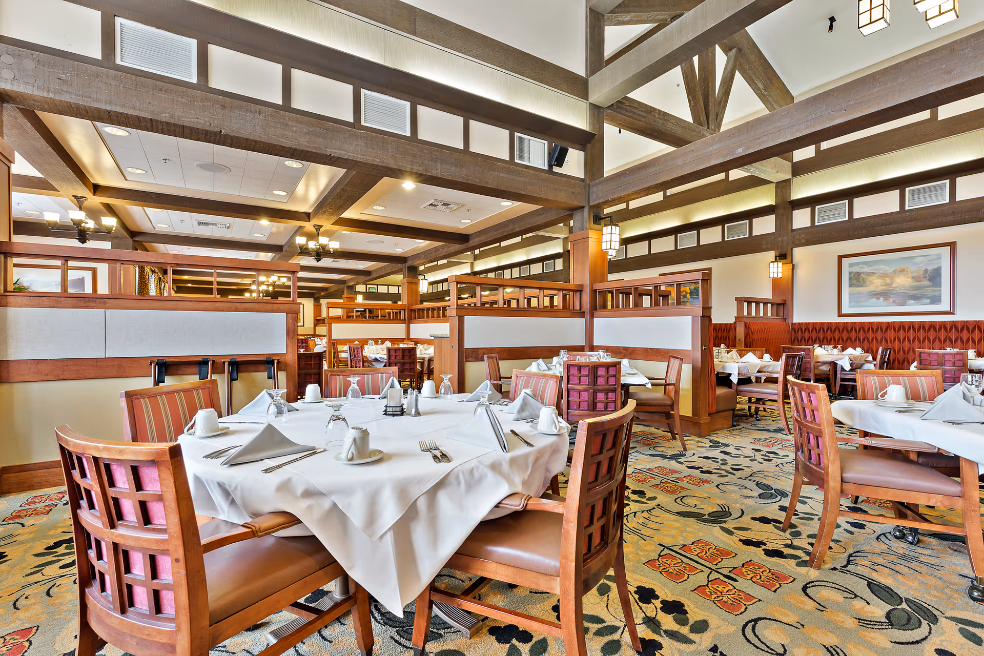 A spacious dining room with wooden beams on the ceiling and multiple tables set with white tablecloths, napkins, cups, and silverware. The room features wooden chairs with cushioned seats, booth seating along the walls, patterned carpet, and warm lighting fixtures. A framed landscape painting is visible on the far wall.