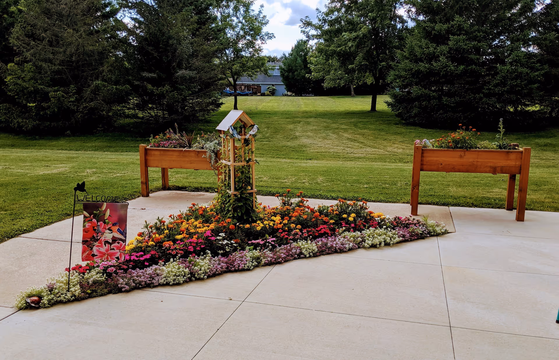 A landscaped outdoor garden area with a triangular flower bed filled with colorful flowers, two raised wooden planters on either side, and a small wooden trellis in the center. The garden is surrounded by a concrete patio and lush green grass with trees in the background under a partly cloudy sky.