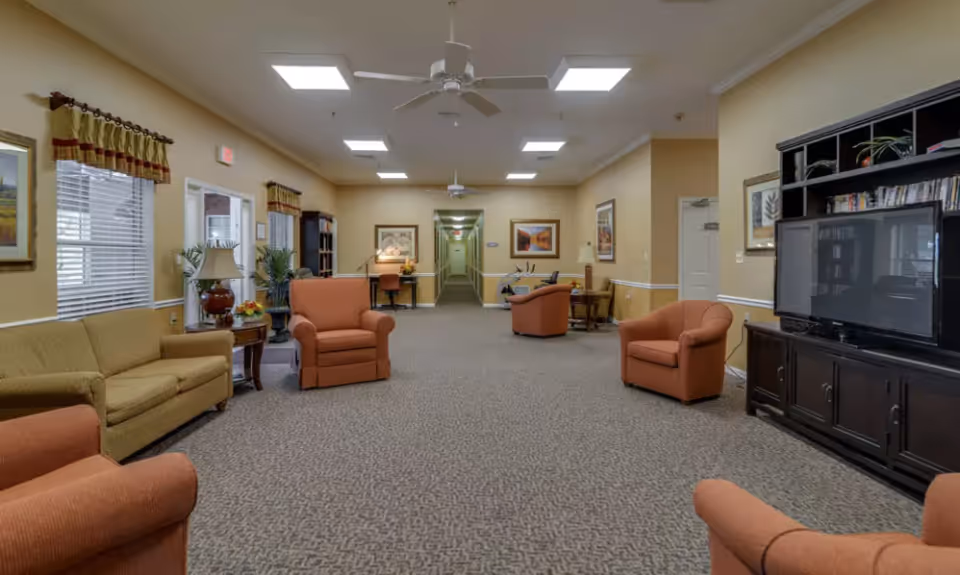 Spacious senior living common room with upholstered chairs and sofas, a TV console, and a long central hallway.