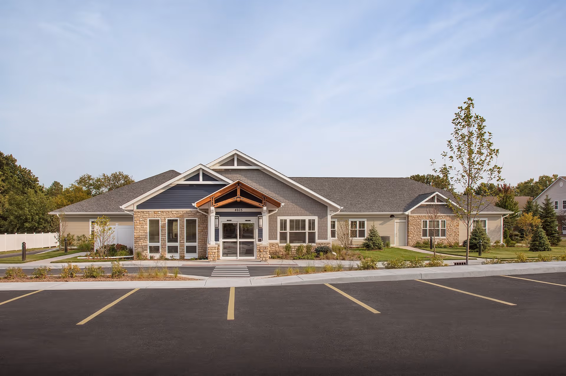 Front exterior view of a single-story senior living facility building with a peaked roof, stone and siding facade, a covered entrance, and a parking lot in front. The building is surrounded by landscaped greenery and trees under a clear sky.