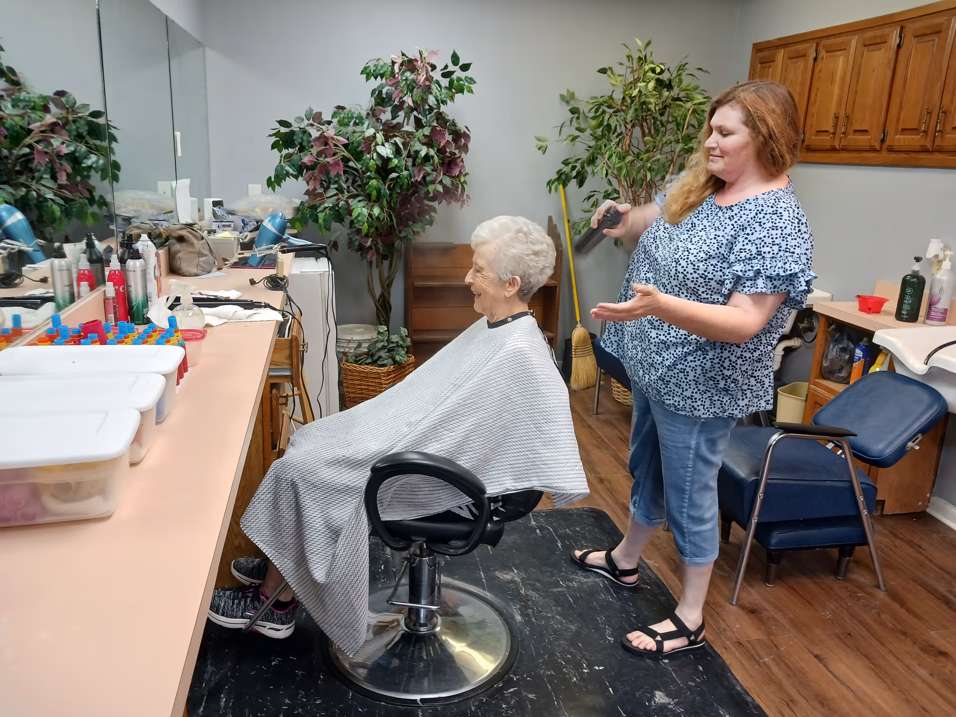 An elderly woman sitting in a salon chair with a white cape around her, smiling while a hairstylist sprays her hair. The room has a large mirror, various hair products on the counter, potted plants, and wooden cabinets in the background.