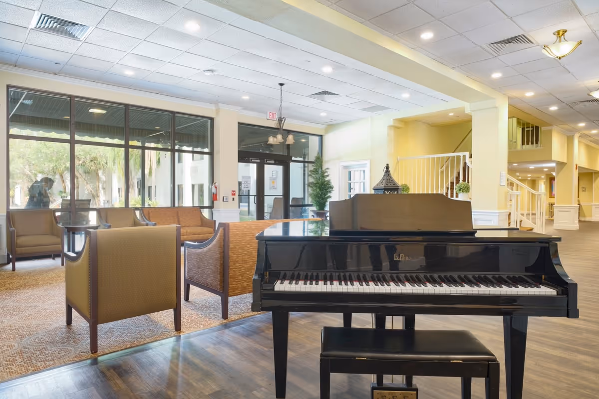 Interior view of a senior living facility lounge area featuring a black grand piano with a matching bench in the foreground. Behind the piano, there are several upholstered chairs and a sofa arranged around a carpeted seating area near large windows letting in natural light. The space has light-colored walls, a tiled ceiling with recessed lighting, and a staircase leading to an upper floor.