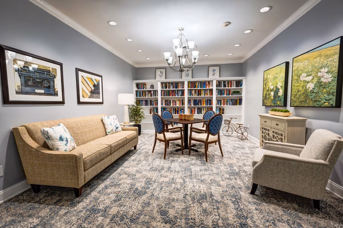 A cozy living room with a beige sofa adorned with floral pillows on the left, a gray armchair on the right, and a round wooden table with four blue cushioned chairs in the center. Behind the table is a large white bookshelf filled with books. The walls are painted light gray and decorated with framed artwork, including a vintage typewriter and floral paintings. A chandelier hangs from the ceiling, and a patterned carpet covers the floor.