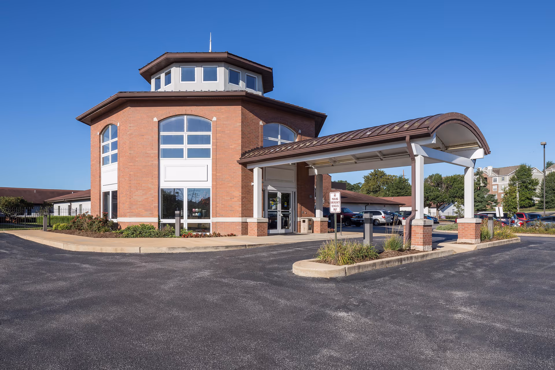 Exterior view of a brick building with large windows and a covered entrance driveway under a clear blue sky. The building is part of a senior care facility with a parking lot and some greenery around.