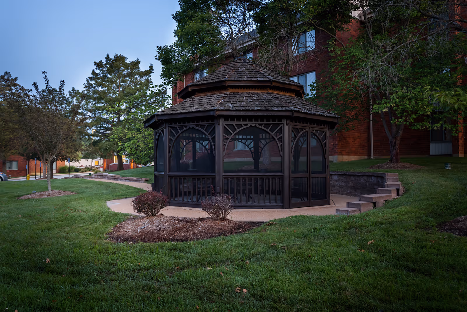 A wooden screened gazebo sits on a grassy lawn in front of a brick senior apartment building.