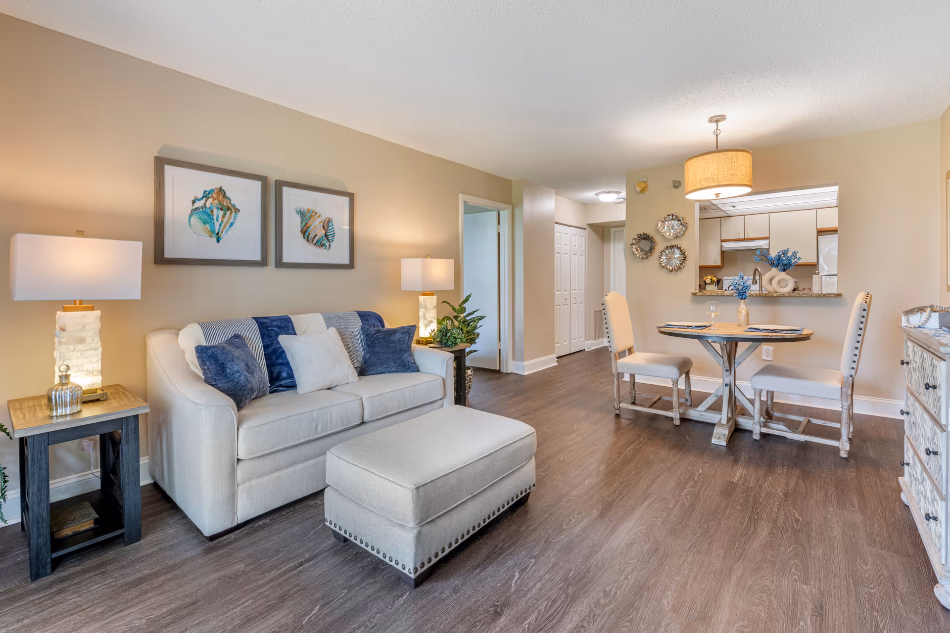 A cozy living room and dining area with a beige sofa adorned with blue and white pillows, a matching ottoman, two side tables with lamps, and two framed seashell artworks on the wall. The dining area features a round wooden table with two upholstered chairs, a hanging light fixture, and a kitchen visible through a pass-through window. The flooring is wood, and the walls are painted beige.