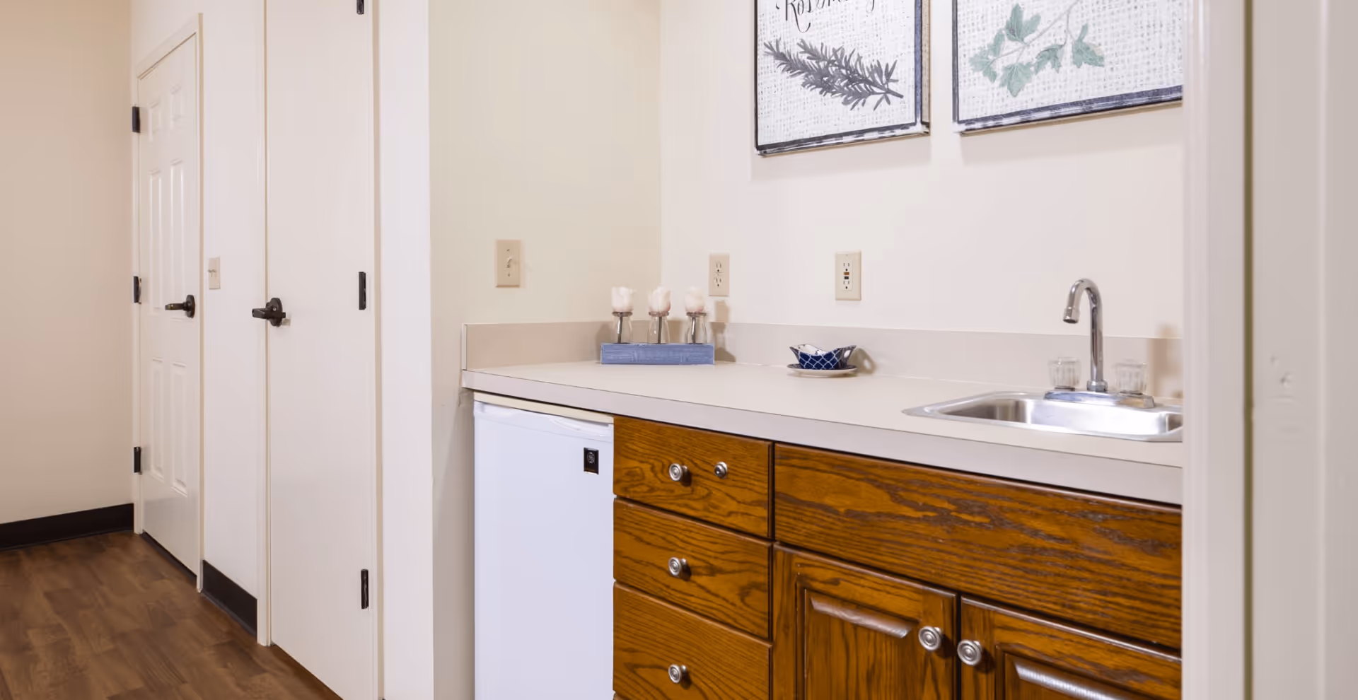 A small kitchenette area with wooden cabinets, a white mini refrigerator, a countertop with a small sink and faucet, and decorative items including three small vases with white flowers and a small blue dish. Two framed botanical prints hang on the wall above the counter. There are two closed white doors to the left and wood flooring.