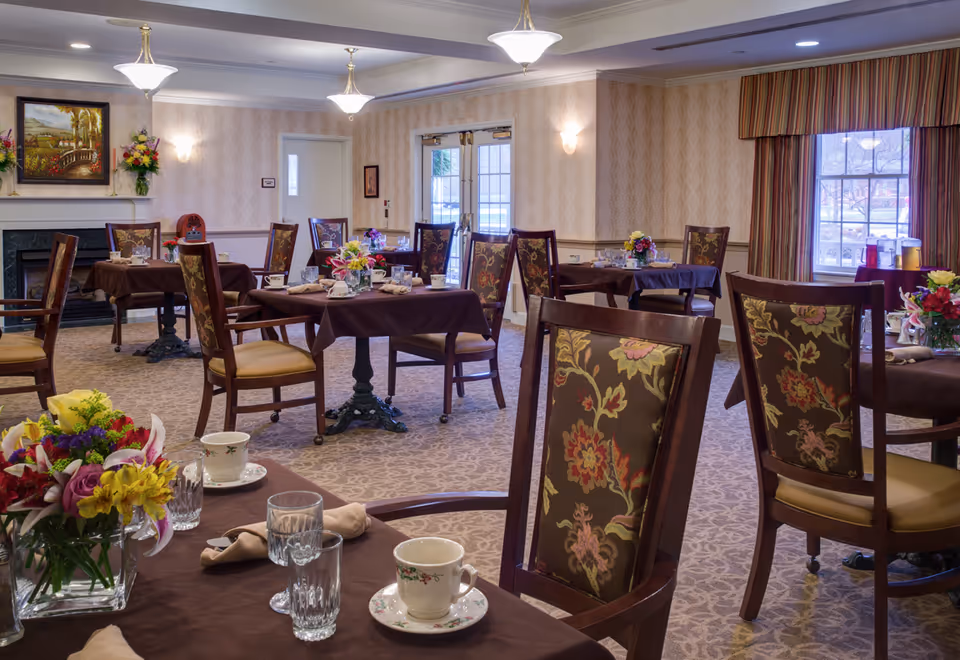 A cozy dining room with several tables covered in brown tablecloths, each set with cups, glasses, napkins, and floral centerpieces. The room has patterned carpet, floral upholstered chairs, soft lighting from ceiling fixtures and wall sconces, a fireplace with a painting above it, and a window with striped curtains letting in natural light.