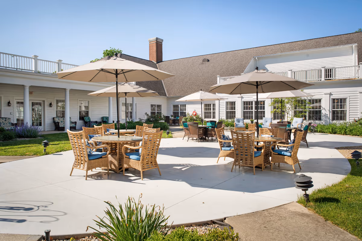 Outdoor patio area at a senior living facility with multiple round tables surrounded by wicker chairs with blue cushions. Large beige umbrellas provide shade over the tables. The patio is surrounded by a white building with multiple windows and a green lawn with plants and shrubs.