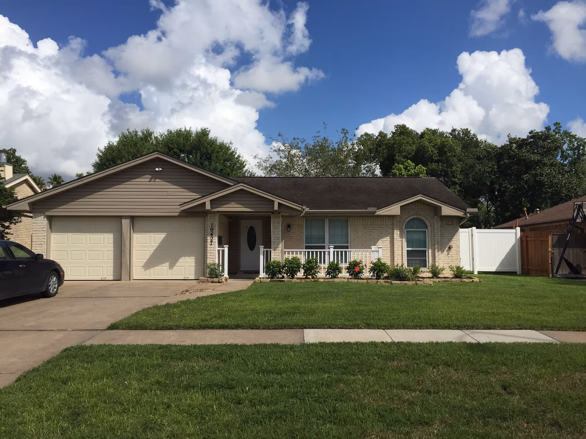 Single-story ranch-style house with an attached two-car garage, front porch, lawn, and driveway under a partly cloudy sky.