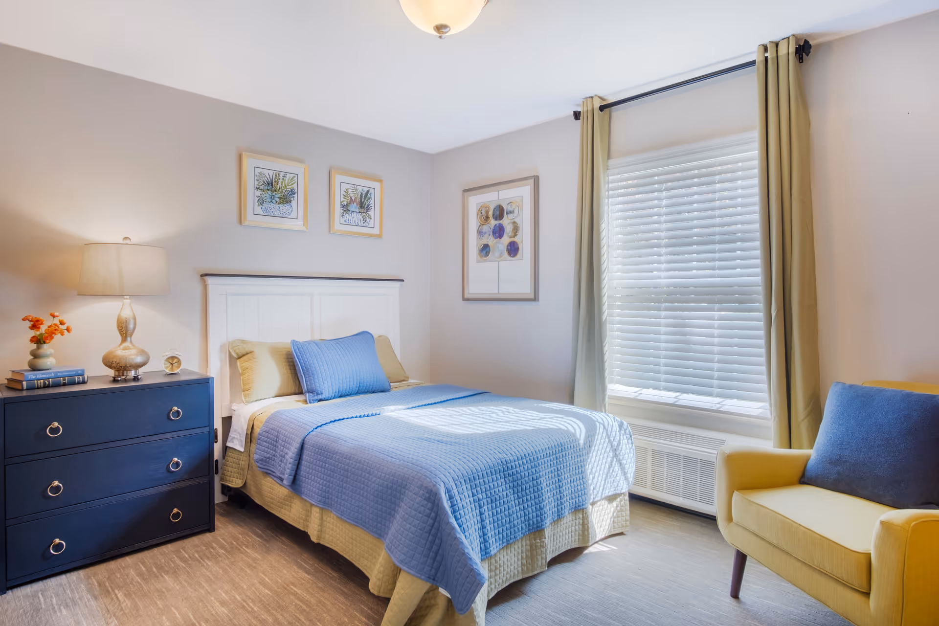A bright and cozy bedroom with a bed covered in blue and beige bedding, a dark blue dresser with a lamp and decorative items, a yellow armchair with a blue cushion, and a window with beige curtains letting in natural light. The walls are decorated with framed artwork.