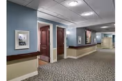 Carpeted interior corridor of a senior living facility with patterned flooring, handrails, framed artwork, recessed ceiling lights, and open apartment doors.