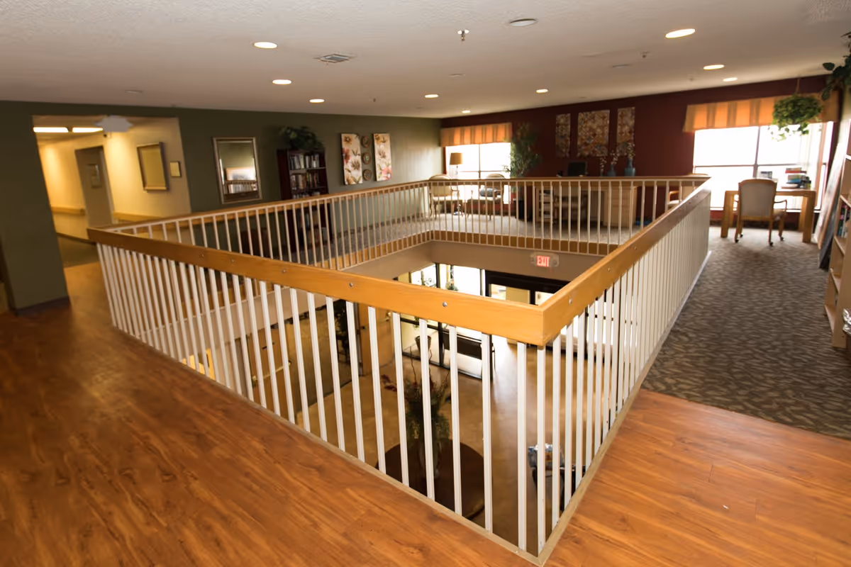 Interior view of a senior living facility showing a second-floor hallway with wooden flooring and white railing overlooking the lobby area below. The hallway leads to a sitting area with chairs, desks, plants, and decorative wall art. The space is well-lit with ceiling lights and natural light from windows.