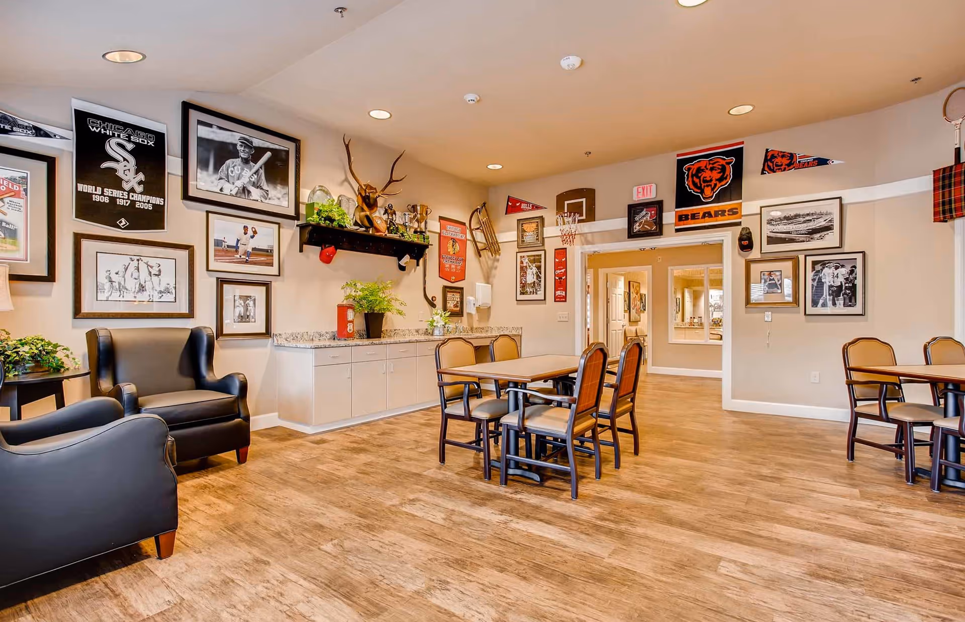 A common area in a senior living facility decorated with Chicago sports memorabilia including Chicago White Sox and Chicago Bears banners and photos. The room has wooden flooring, beige walls, two black armchairs, tables with chairs, and a countertop with plants and trophies.