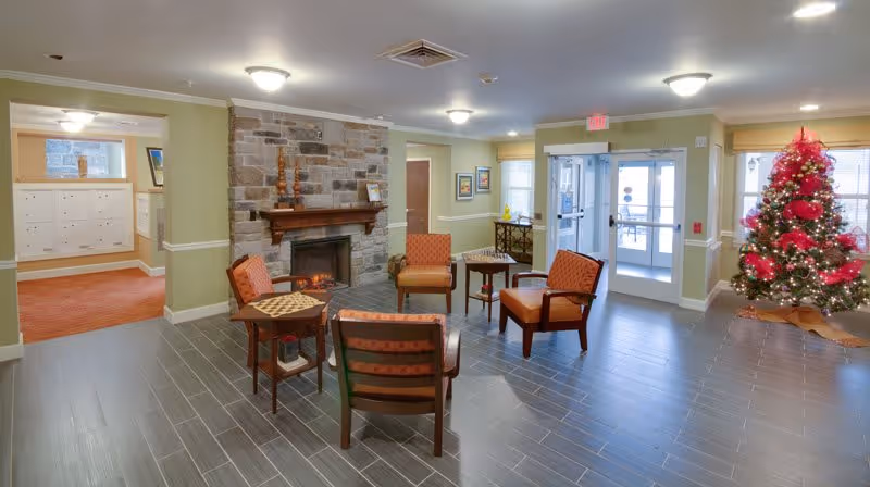 Bright lobby-style common room with chairs arranged around a stone fireplace and a decorated Christmas tree by glass entrance doors.