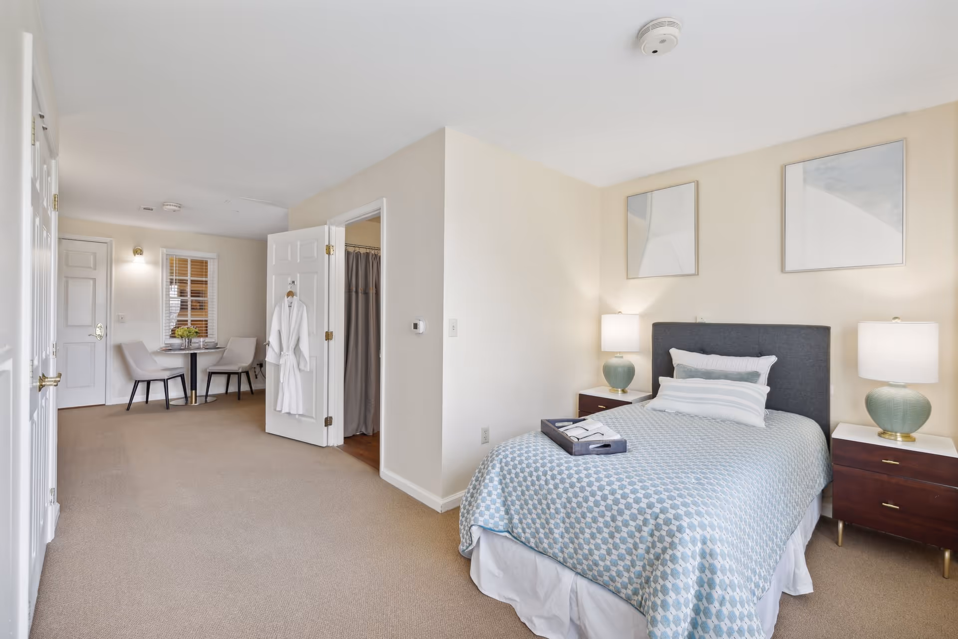 A bright and clean bedroom in a senior living facility with a neatly made bed featuring a blue patterned bedspread and multiple pillows. Two matching bedside tables with green lamps flank the bed. Two abstract framed artworks hang above the bed. To the left, an open door reveals a bathroom with a shower curtain. Further left, a small dining area with a round table and two chairs is visible near a window with blinds. The room has beige walls and carpeted floors.