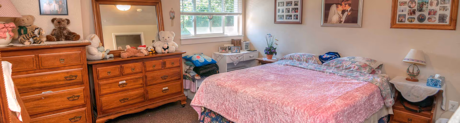 A cozy bedroom with a double bed covered in a pink bedspread and floral pillows. There are wooden dressers with stuffed animals on top, a mirror, and framed pictures on the walls. A window with blinds lets in natural light, and there is a small table with a lamp and tissue box beside the bed.