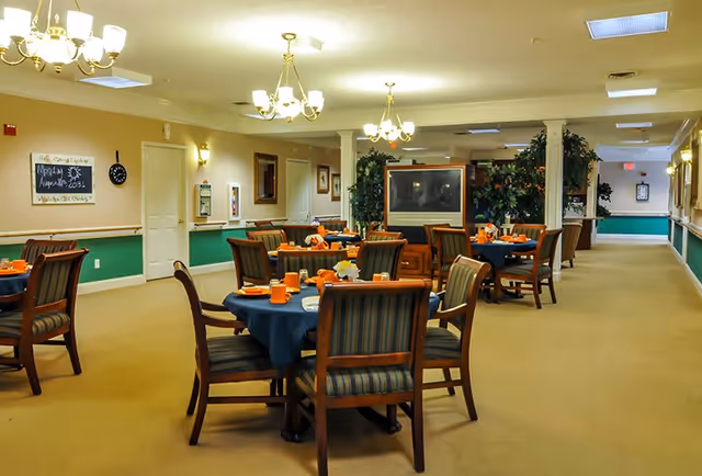 A dining room with several round tables covered with dark blue tablecloths, each set with orange cups and small floral centerpieces. Wooden chairs with striped cushions surround the tables. The room has beige walls with green wainscoting, chandeliers hanging from the ceiling, and a large television set against a wall with plants around it. There is a hallway extending to the right side of the room.