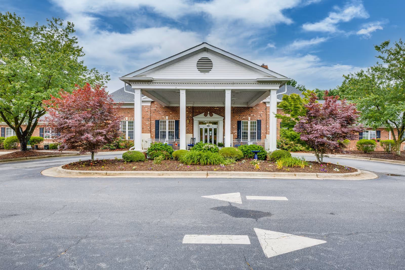 Front exterior view of Kerner Ridge Assisted Living building with a covered entrance supported by white columns, surrounded by landscaped greenery and trees under a partly cloudy sky.