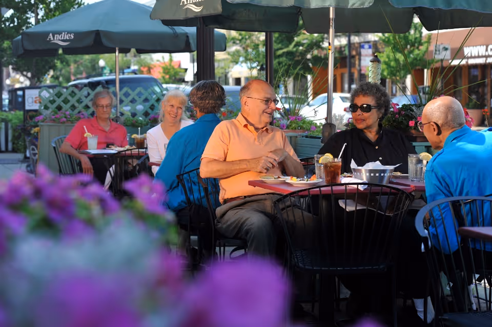 A group of elderly people sitting and chatting at outdoor tables under umbrellas at a restaurant or cafe, with flowers in the foreground and greenery around.