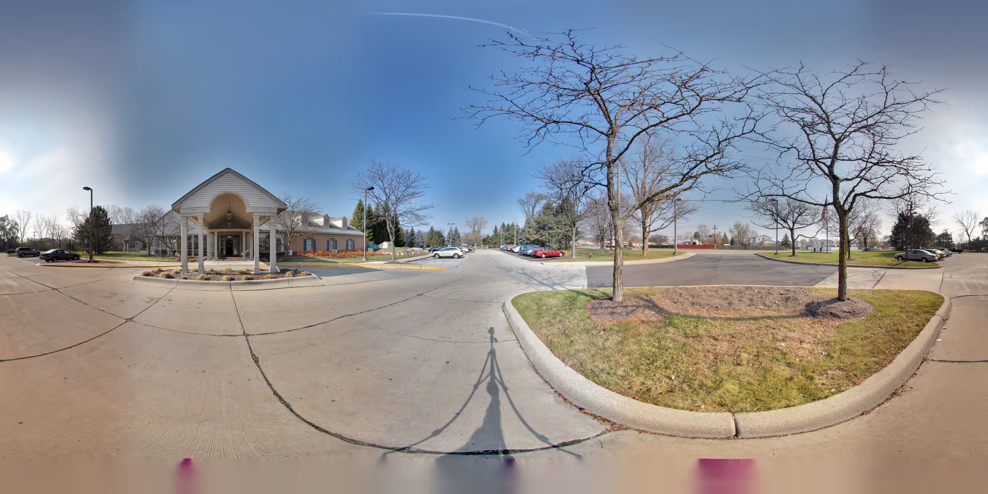 Wide panoramic view of the parking lot and entrance area of Oakmont Livonia Senior Community. The image shows a covered entrance with white pillars, several parked cars, leafless trees, and a clear blue sky.