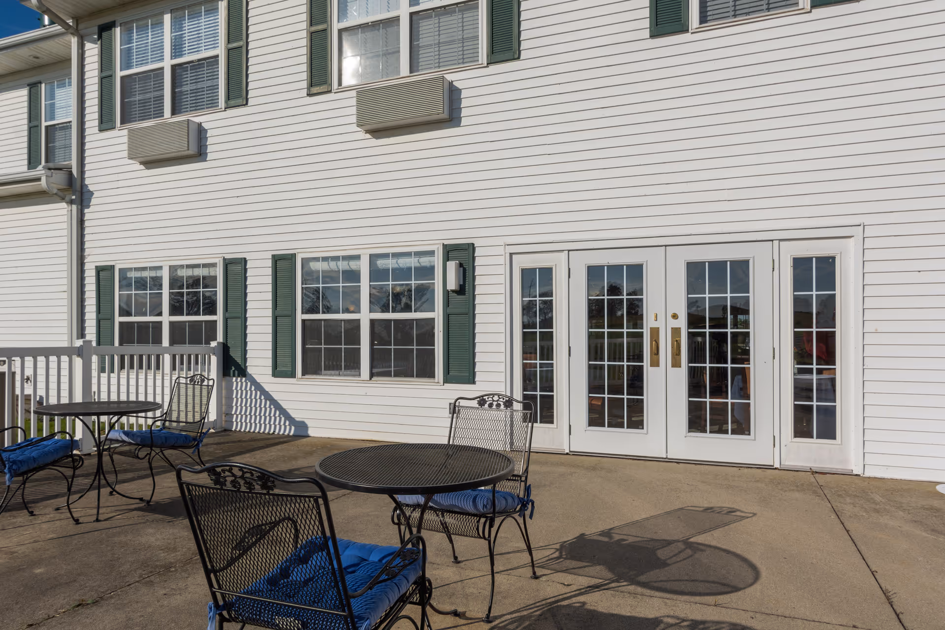 Outdoor patio area with black metal tables and chairs with blue cushions, adjacent to a white building with green window shutters and glass double doors.
