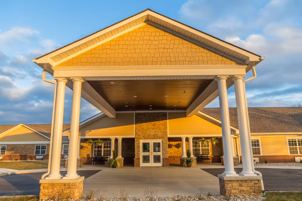 Front exterior view of The Willows at Fritz Farm building with a large covered entrance supported by white columns, brick and yellow siding, and a clear sky with some clouds.