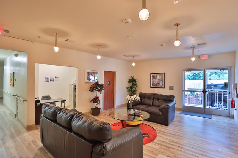 A bright and welcoming living room area in an assisted living facility with two dark leather sofas arranged around a glass coffee table on a red circular rug. The room has light wood flooring, several hanging ceiling lights, framed artwork on the walls, potted plants, and a glass door leading outside with a view of a parked car.