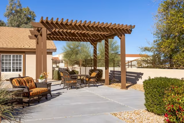 Outdoor patio area with a wooden pergola providing partial shade over a table and four chairs. There is additional seating with cushioned chairs on the concrete patio surrounded by desert landscaping including bushes, rocks, and trees. The sky is clear and blue.