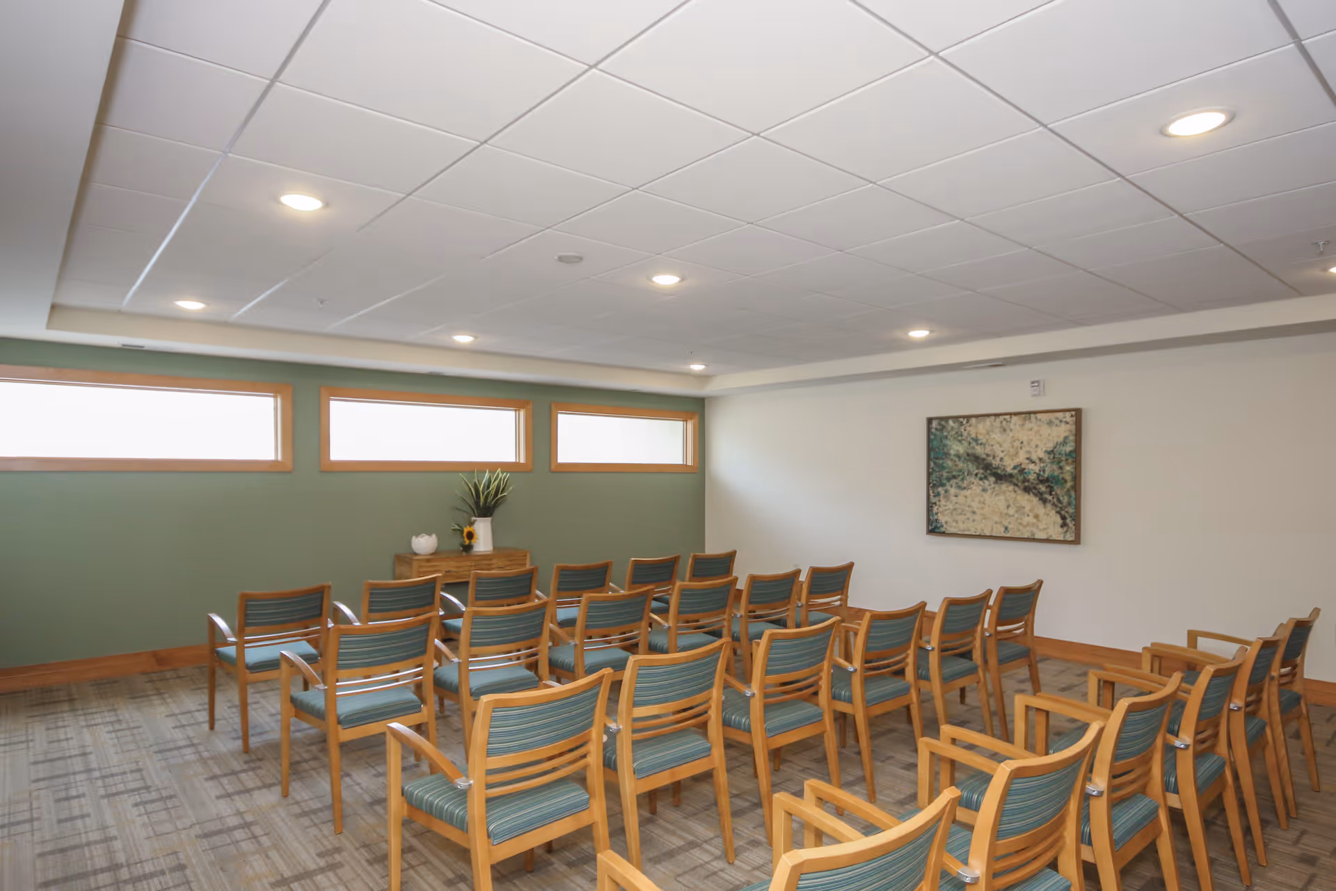 A small meeting or activity room with rows of wooden chairs featuring green cushions, a green accent wall with three rectangular windows, a small wooden table with a plant and decorative items, and a framed abstract painting on a white wall.