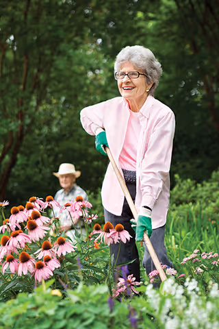 An elderly woman wearing glasses, a pink shirt, and green gardening gloves is smiling while gardening with a hoe among blooming pink flowers. In the background, an elderly man wearing a hat is sitting and watching her in a lush green garden.